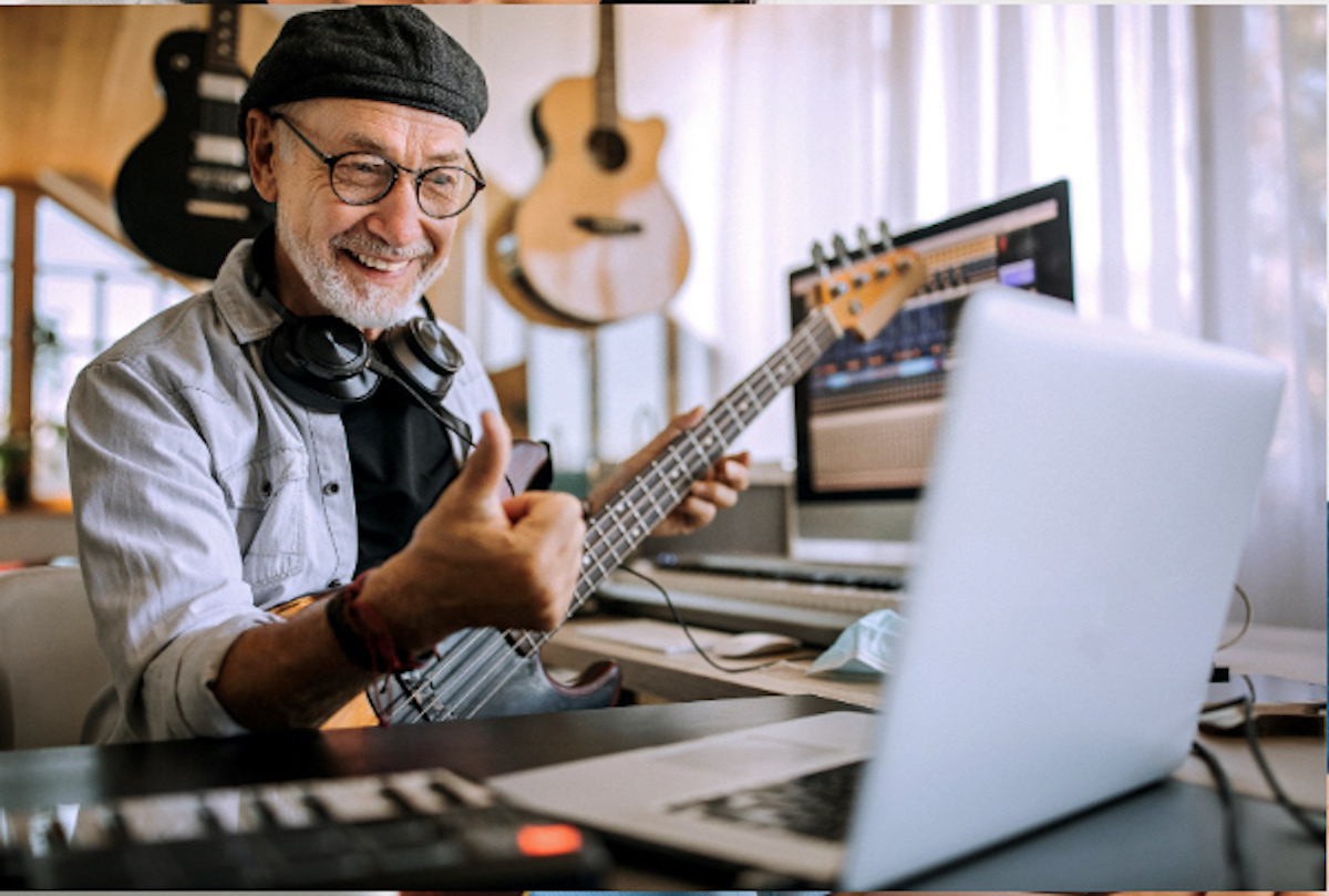 Smilende mand spiller basguitar under en online musiklektion og giver tommel op til kameraet.